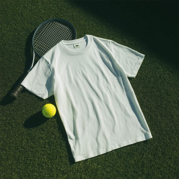 A mint green t-shirt lies flat on a tennis court, next to a tennis racket and a yellow tennis ball