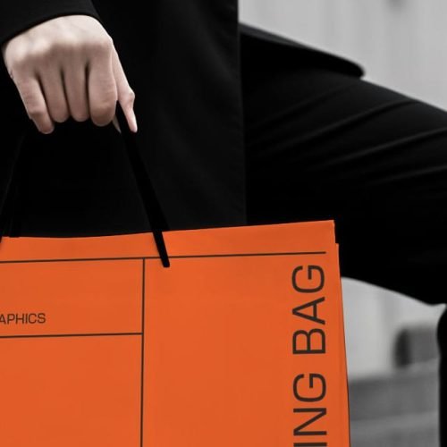 A woman in a black suit and heels holds an orange shopping bag labeled "FREE MOCKUPS" while walking up stairs